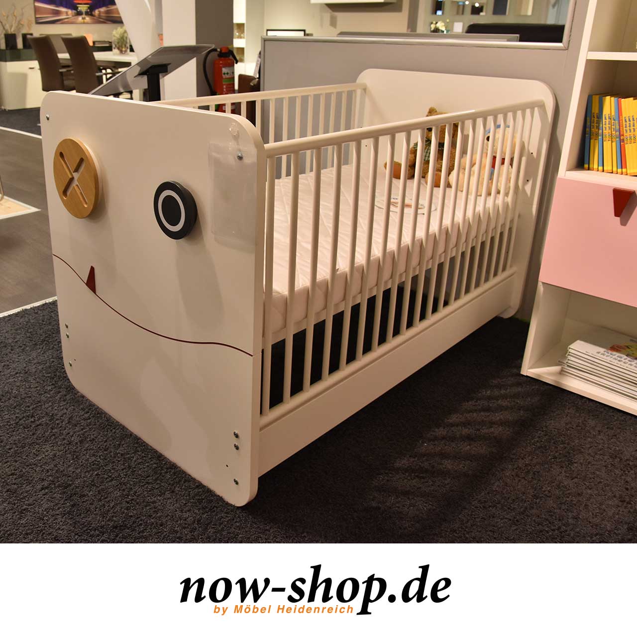 Weißes Babybett mit Sprossen an der Seite. An der Front zwei Haken, die wir Augen aussehen und ein angedeuteter Mund mit rotem Zähnchen.